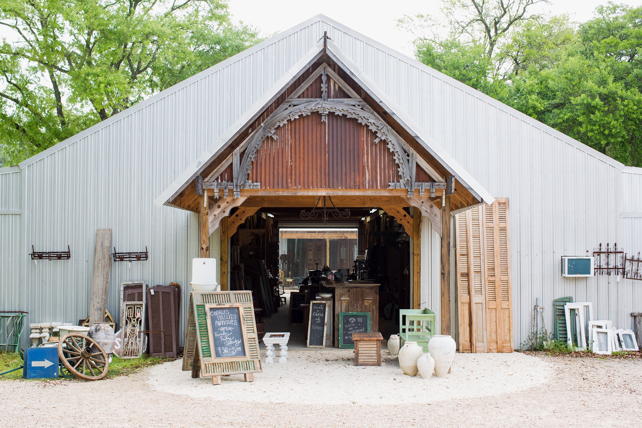 Vintage-style antique shop with open barn door, surrounded by outdoor decor and furniture, with trees in the background.