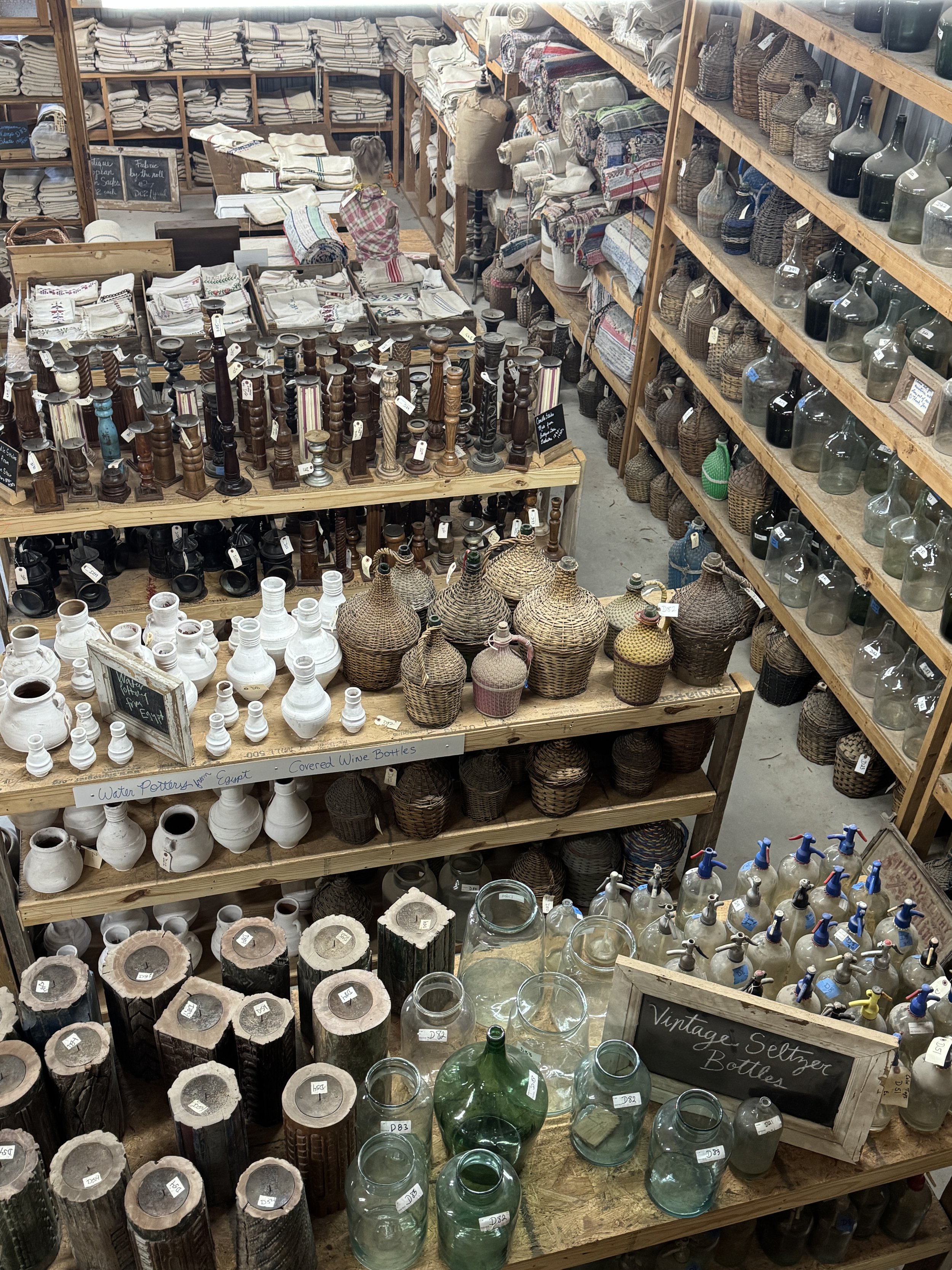 Shelves filled with various vintage glass bottles, ceramic jars, and woven baskets, with some items labeled with prices in a rustic store.