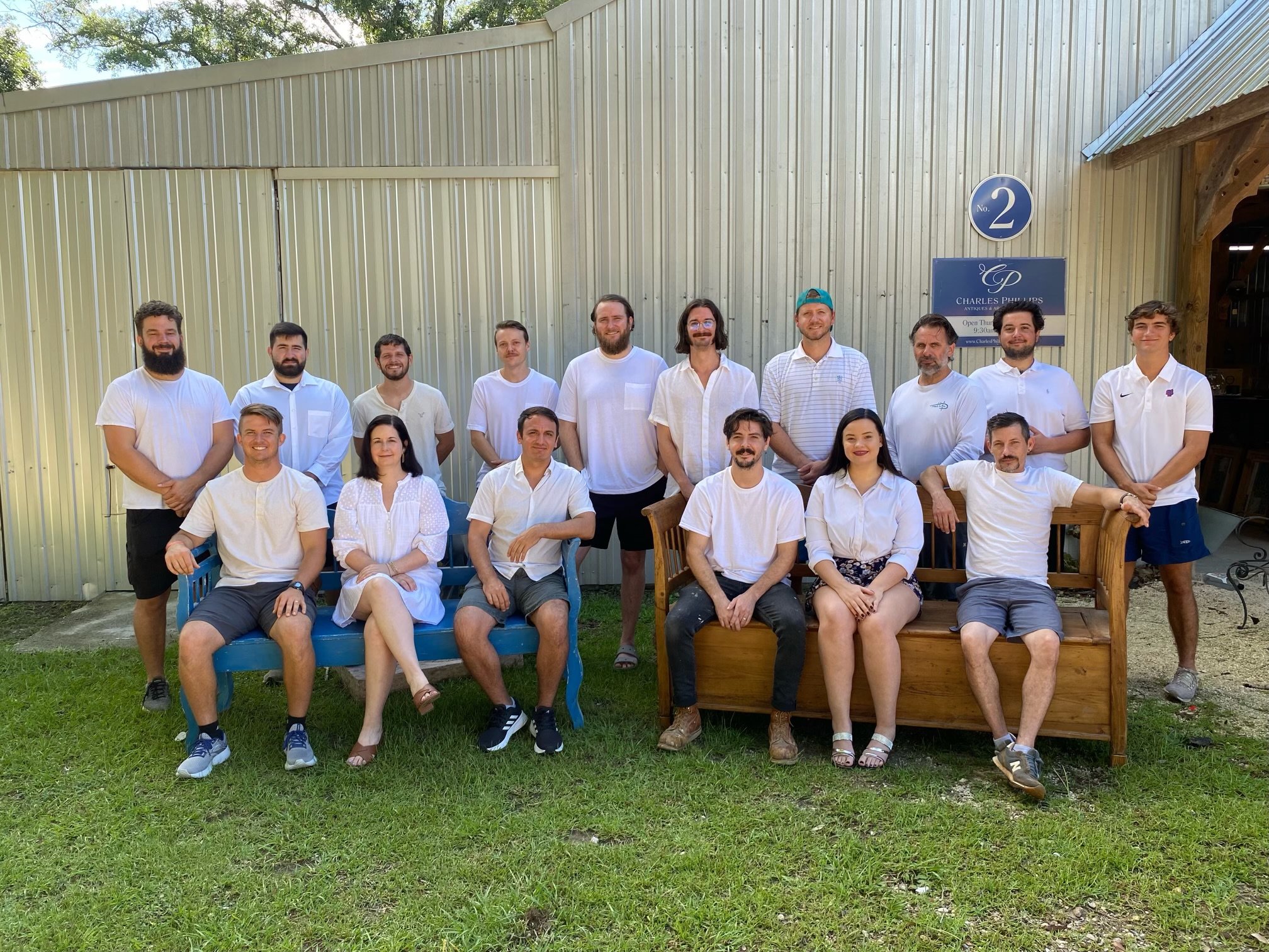 Group of fifteen people, mostly young adults, posing outdoors in front of a siding building. Some are sitting on a bench, others standing. They are casually dressed, many in white shirts and shorts or skirts, with a mix of smiling and relaxed expressions.