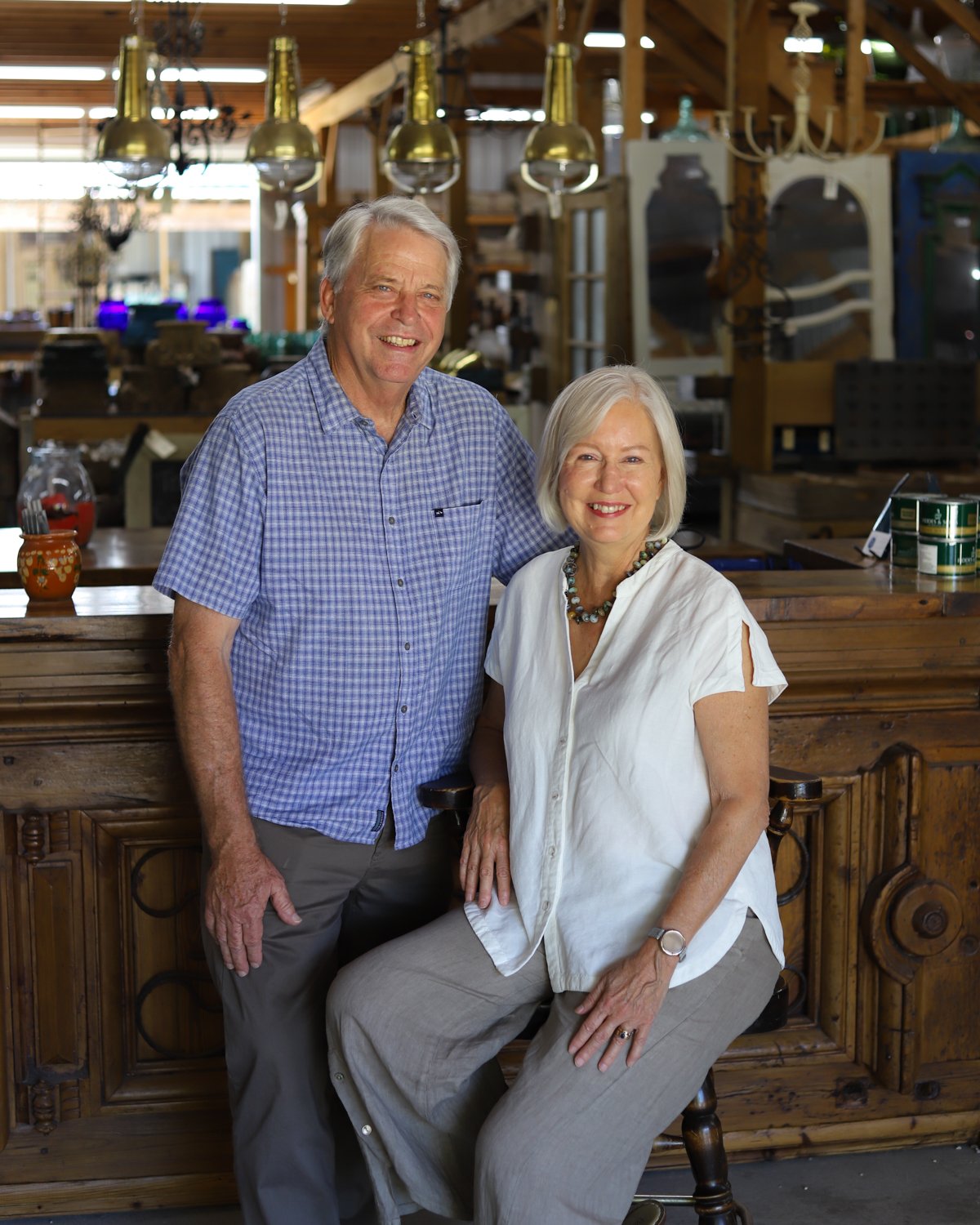 An elderly couple smiling, the man standing and the woman seated, in a rustic wooden interior, possibly a shop or café.