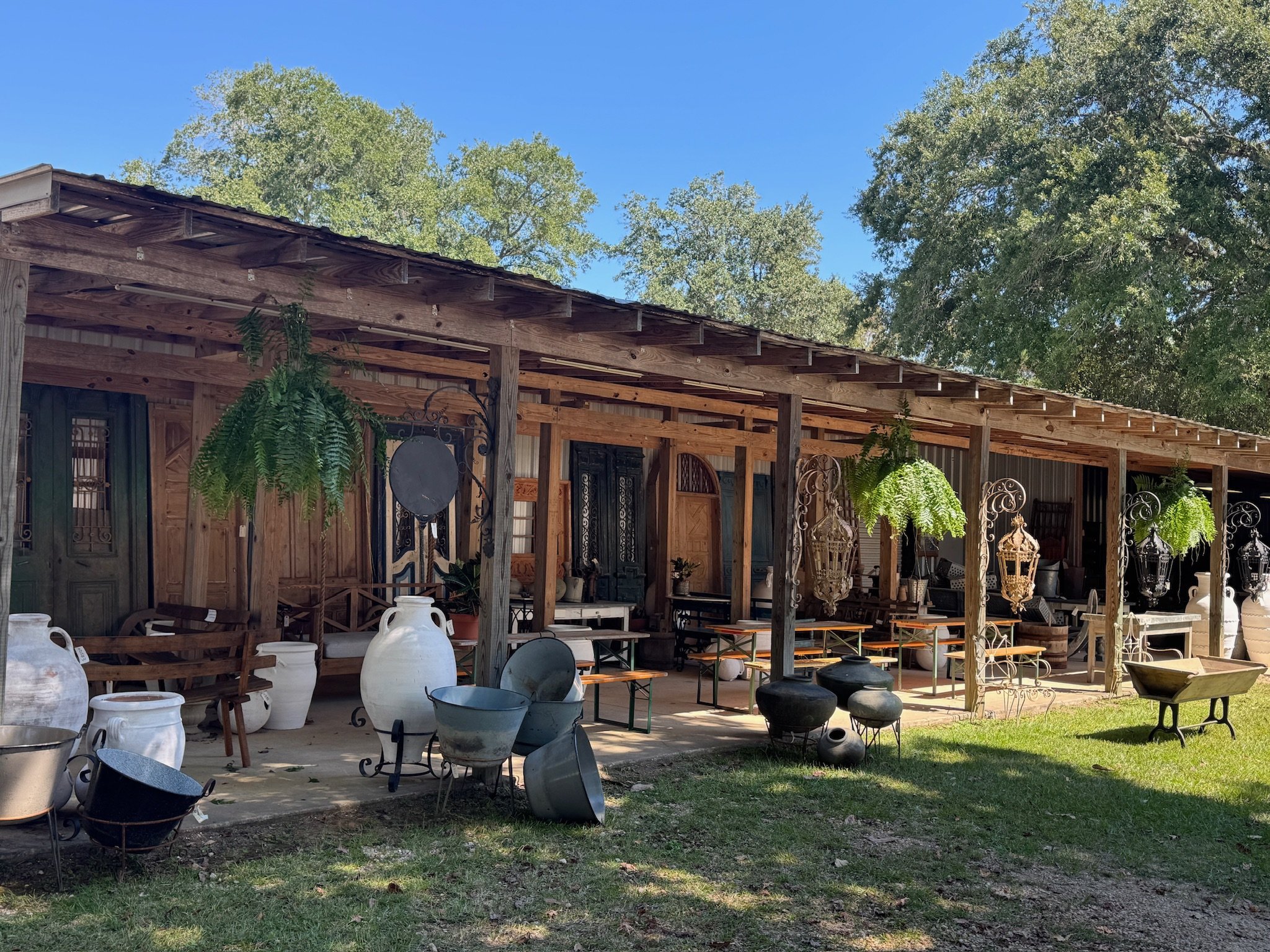 Outdoor display of vintage and antique pottery, metal buckets, and decorative items in front of a wooden shed with green ferns hanging from the beams.
