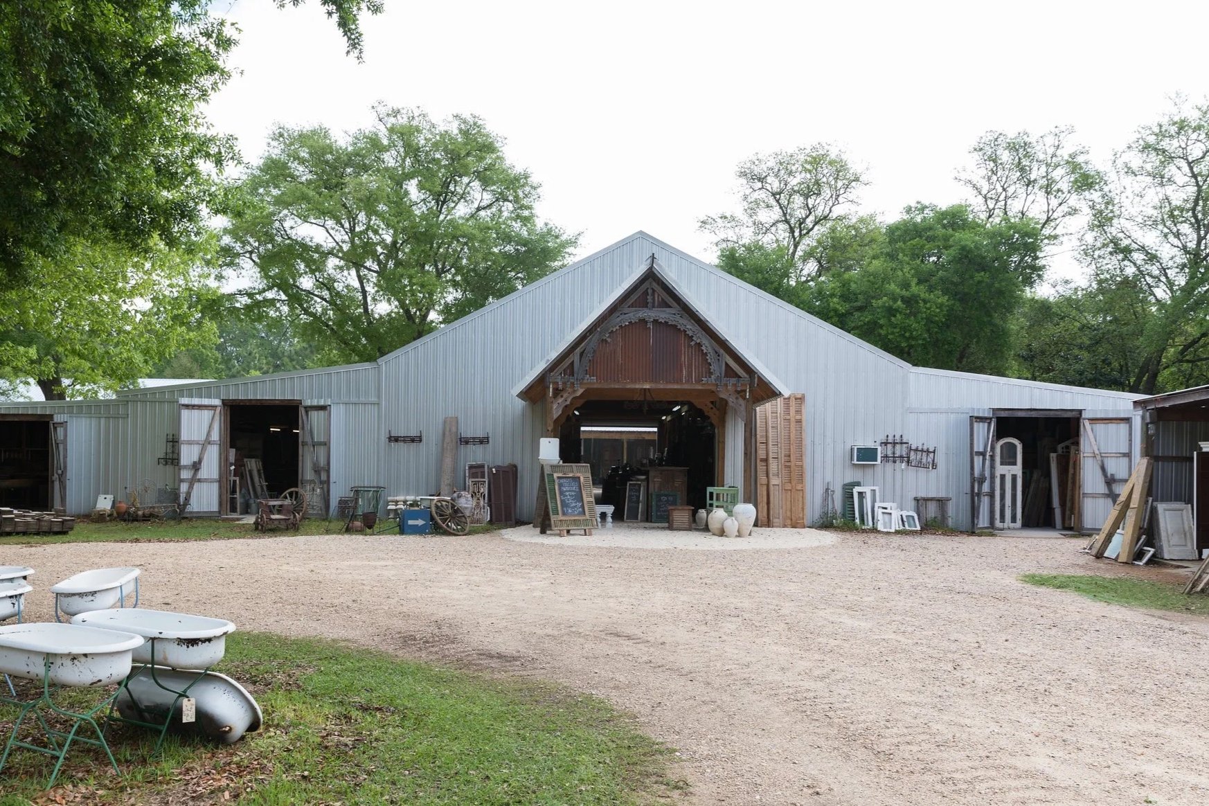View of a rustic metal barn with open doors, displaying various vintage and antique items outside, surrounded by green trees.