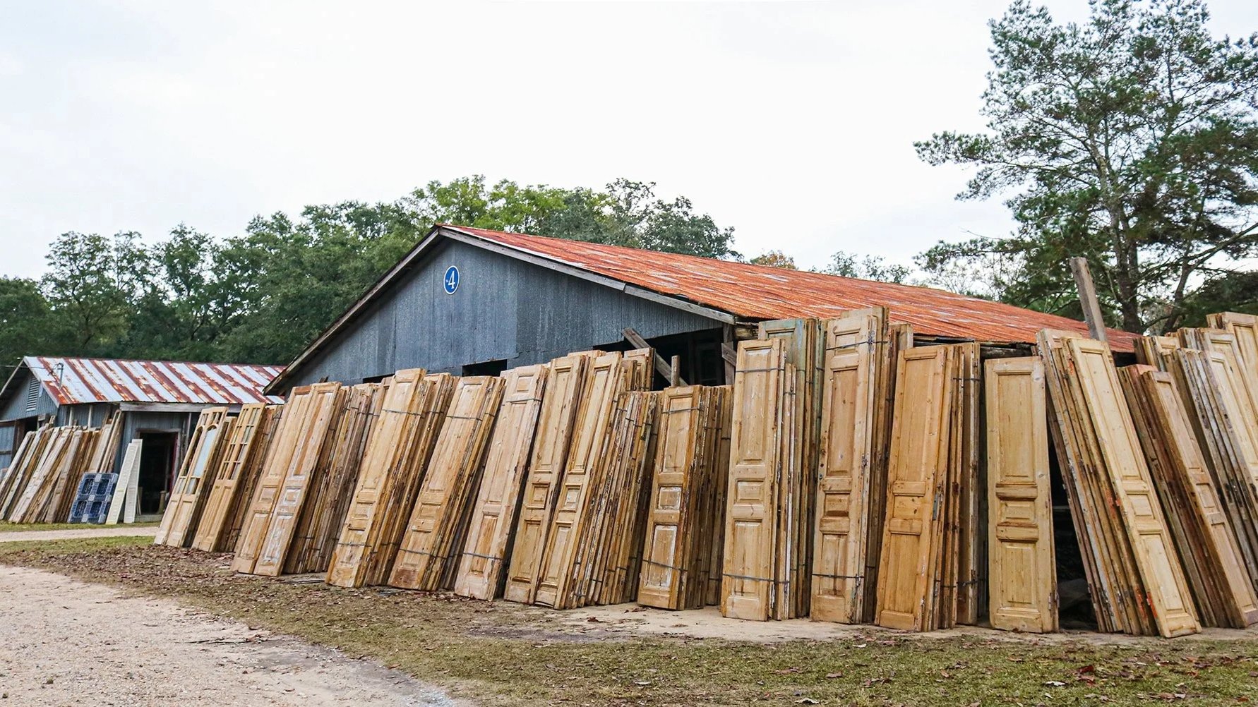 Old wooden doors leaning against a rustic building with a rusty metal roof, set in a rural outdoor area with trees in the background.