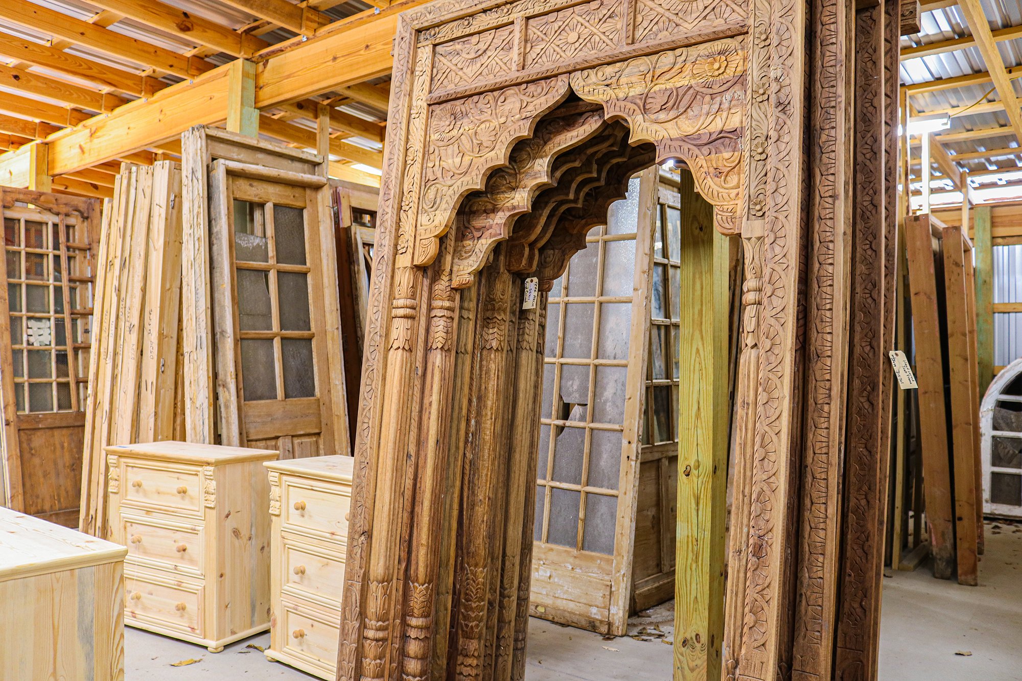A woodworking shop with various wooden doors, including an ornate carved doorway, and two wood dressers in the foreground.